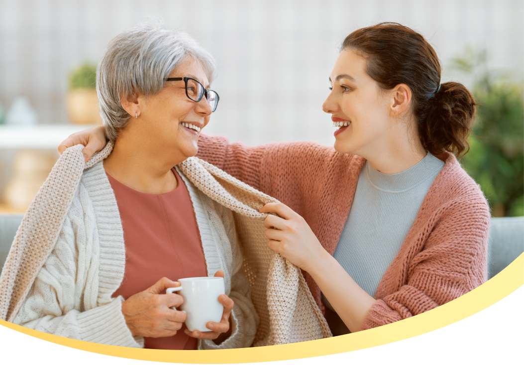 A woman smiles wrapping a blanket around another woman, who holds a cup of coffee.